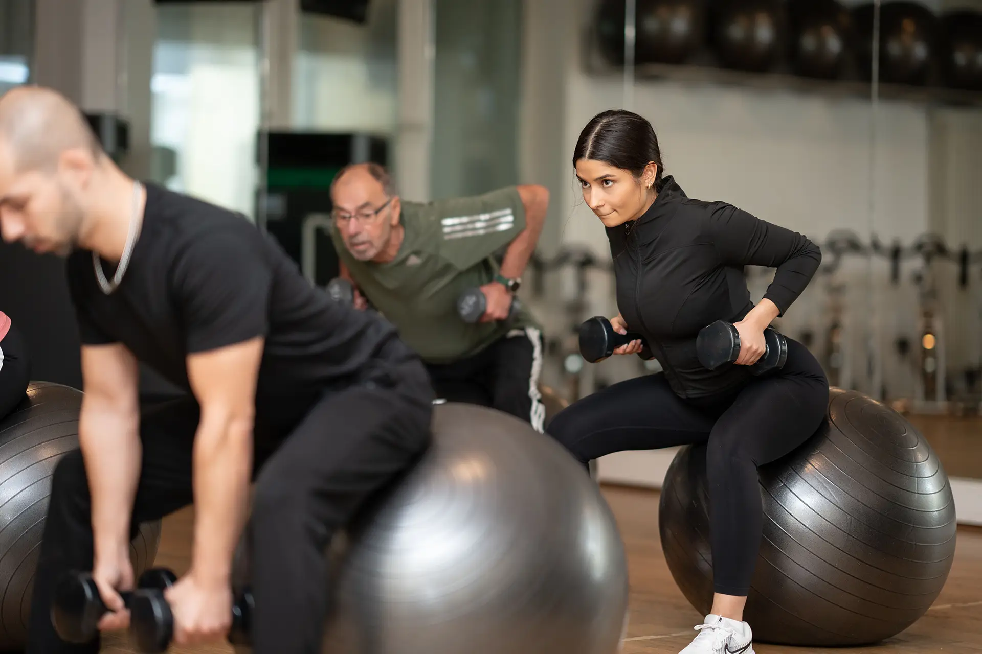 Teilnehmende sitzen auf Gymnastikbällen und führen mit Kurzhanteln eine Ruderbewegung aus. Im Fokus ist eine Frau in schwarzer Sportkleidung, die konzentriert nach hinten zieht, während weitere Teilnehmende die Übung im Hintergrund mitmachen. Der Raum ist hell und mit Spiegeln und Fitnessgeräten ausgestattet.