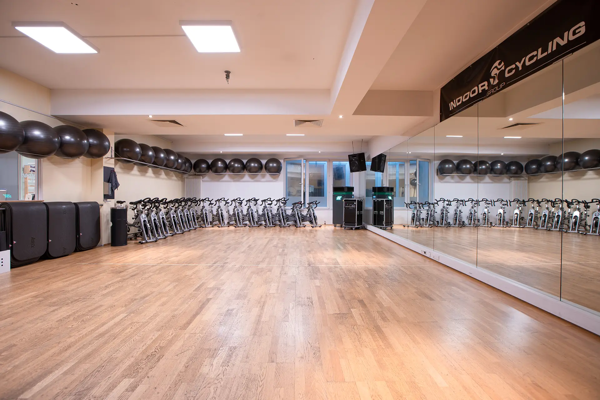Leerer Kursraum mit hellem Holzboden, einer großen Spiegelwand und einer langen Reihe von Indoor-Cycling-Bikes entlang der Rückwand. An der Decke und an der Wand sind schwarze Gymnastikbälle gelagert. Der Raum ist hell ausgeleuchtet und wirkt offen, aufgeräumt und bereit für den nächsten Kurs.
