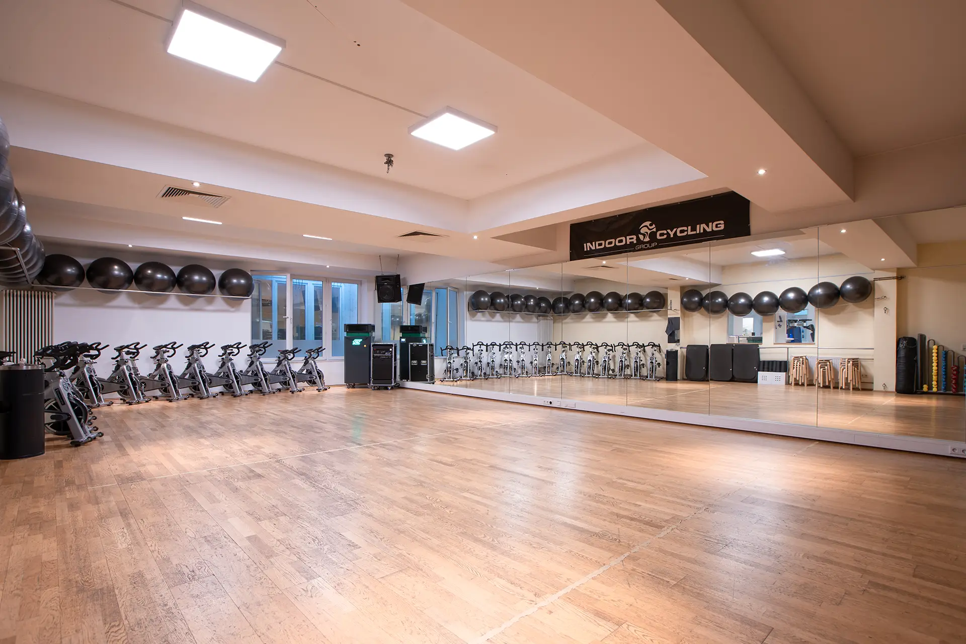 Leerer Kursraum mit hellem Holzboden, einer großen Spiegelwand und einer langen Reihe von Indoor-Cycling-Bikes entlang der Rückwand. An der Decke und an der Wand sind schwarze Gymnastikbälle gelagert. Der Raum ist hell ausgeleuchtet und wirkt offen, aufgeräumt und bereit für den nächsten Kurs.