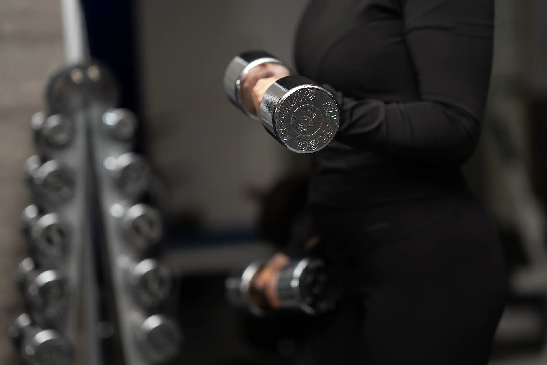 Person führt eine Kraftübung mit Kurzhanteln im Fitnessstudio body & soul Billerbeck aus, Hantelablage im Hintergrund