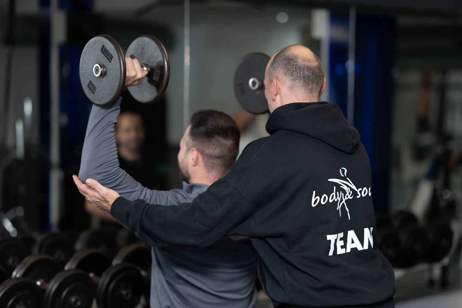 Trainer unterstützt einen Mann beim Schulterdrücken mit Kurzhanteln vor einem Spiegel im Fitnessstudio, Fokus auf Technik, Sicherheit und betreutes Krafttraining