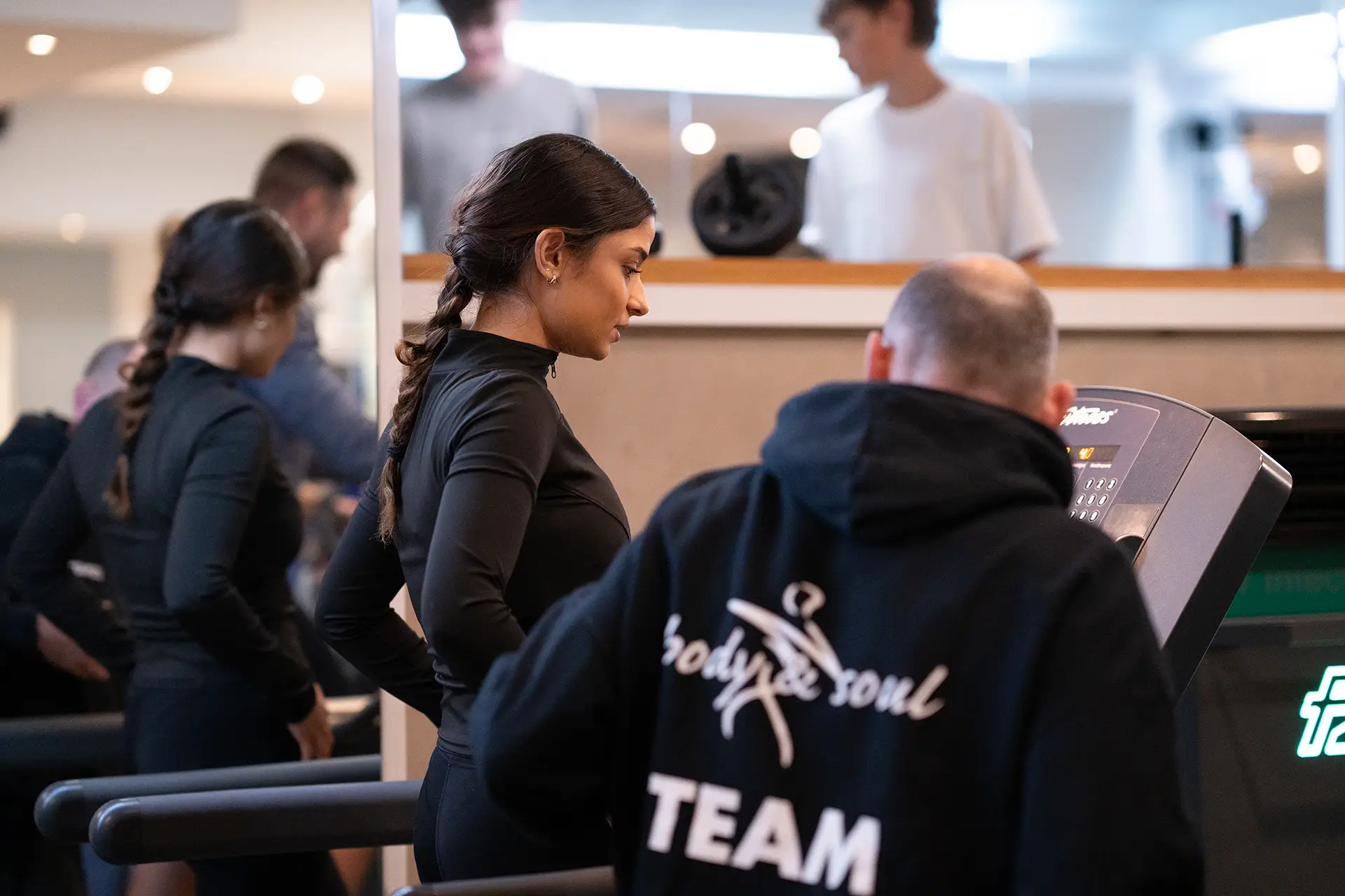 Trainer unterstützt eine Frau bei der Einstellung eines Laufbandes im Fitnessstudio body & soul Billerbeck, weitere Mitglieder trainieren im Hintergrund