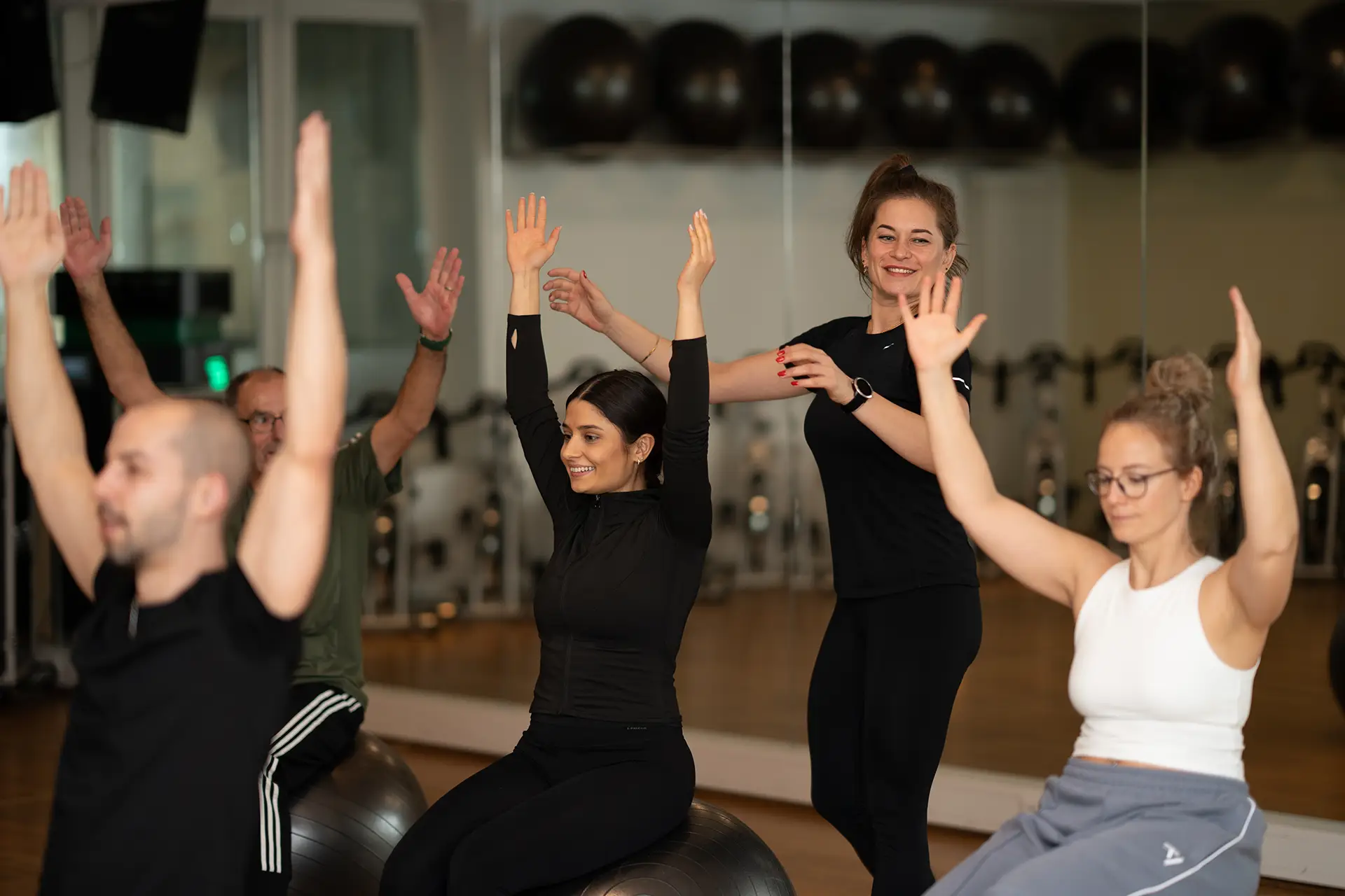 Trainerin unterstützt eine Gruppe von Teilnehmenden, die auf Gymnastikbällen sitzen und mit erhobenen Armen eine Übung ausführen. Alle sind konzentriert und lächeln in einem hellen Trainingsraum mit Spiegelwand im Hintergrund.