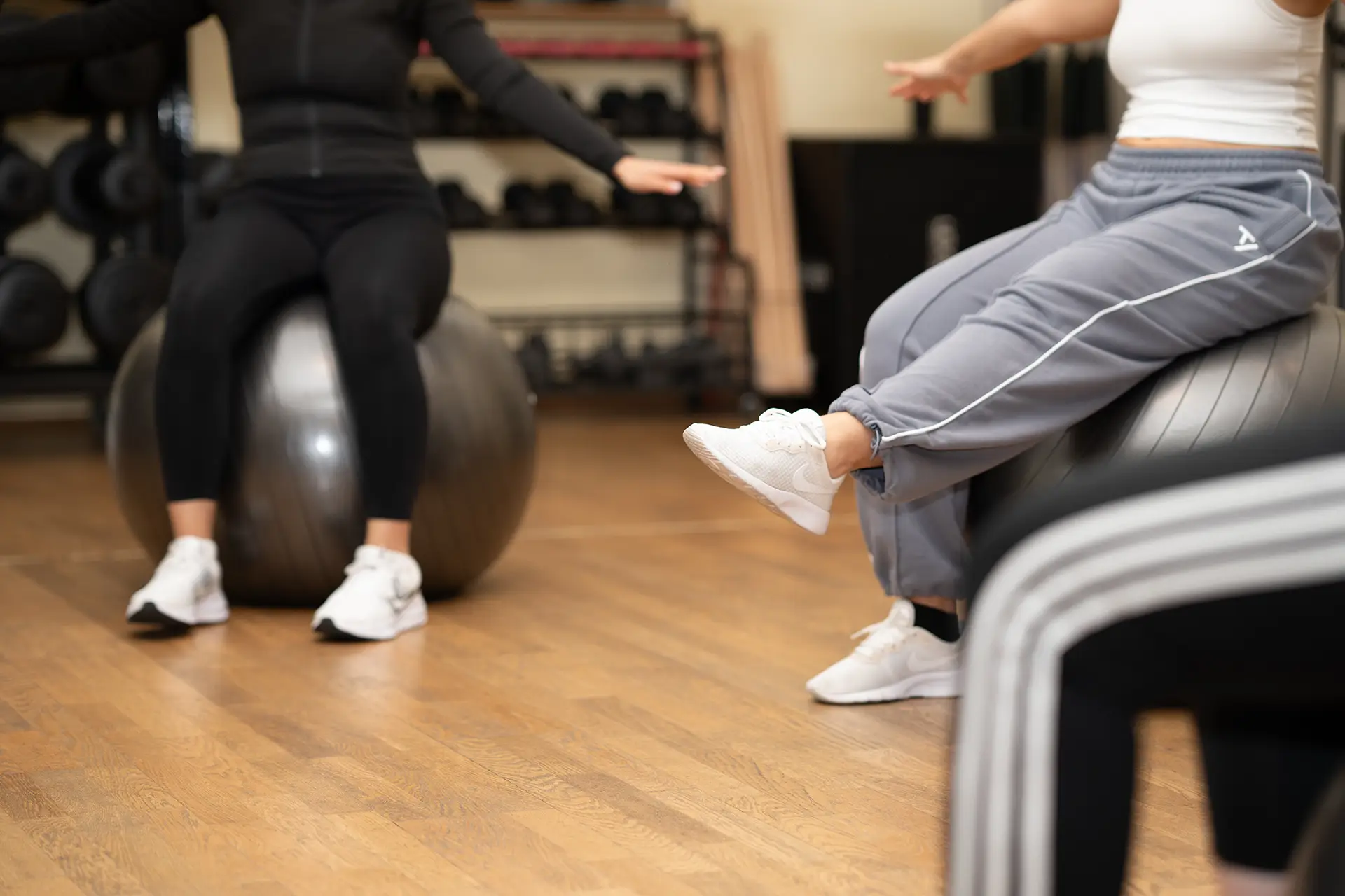 Mehrere Personen sitzen auf Gymnastikbällen im Fitnessstudio und strecken jeweils ein Bein nach vorne, während ihre Sportschuhe, der Holzboden und weitere Trainingsbälle im Hintergrund sichtbar sind.