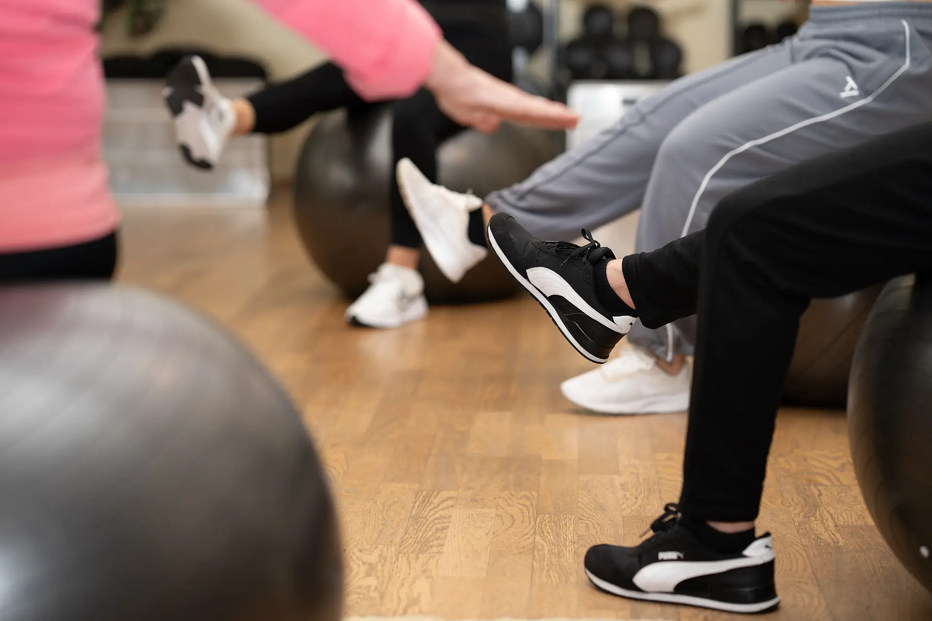 Mehrere Personen sitzen auf Gymnastikbällen im Fitnessstudio und heben ein Bein nach vorne an, während ihre Füße und Sportschuhe im Fokus stehen und der Holzboden sowie weitere Trainingsbälle im Hintergrund zu sehen sind.