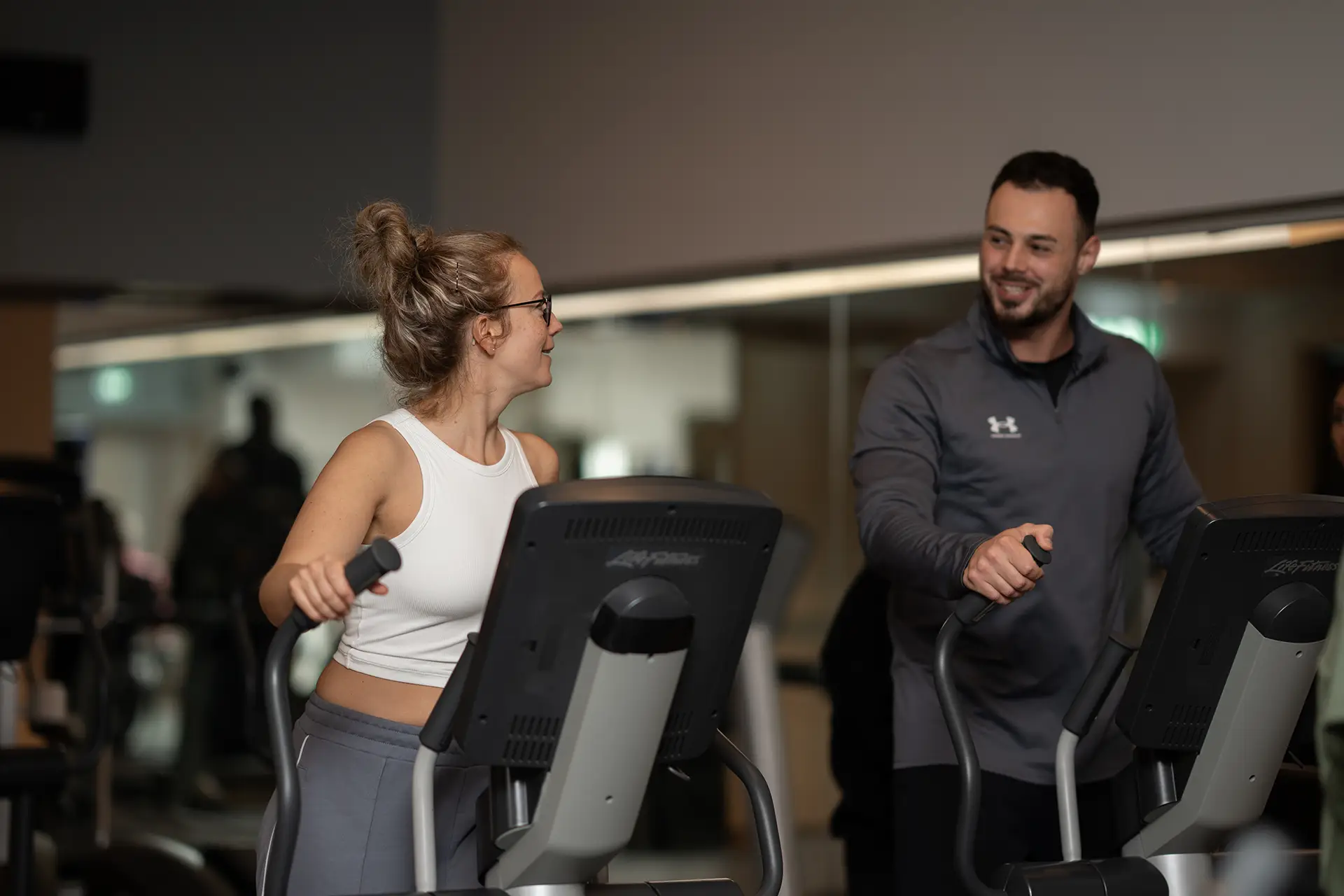 Trainer spricht mit einer Frau während des Trainings an einem Crosstrainer im Fitnessstudio body & soul Billerbeck, beide lächeln sich an