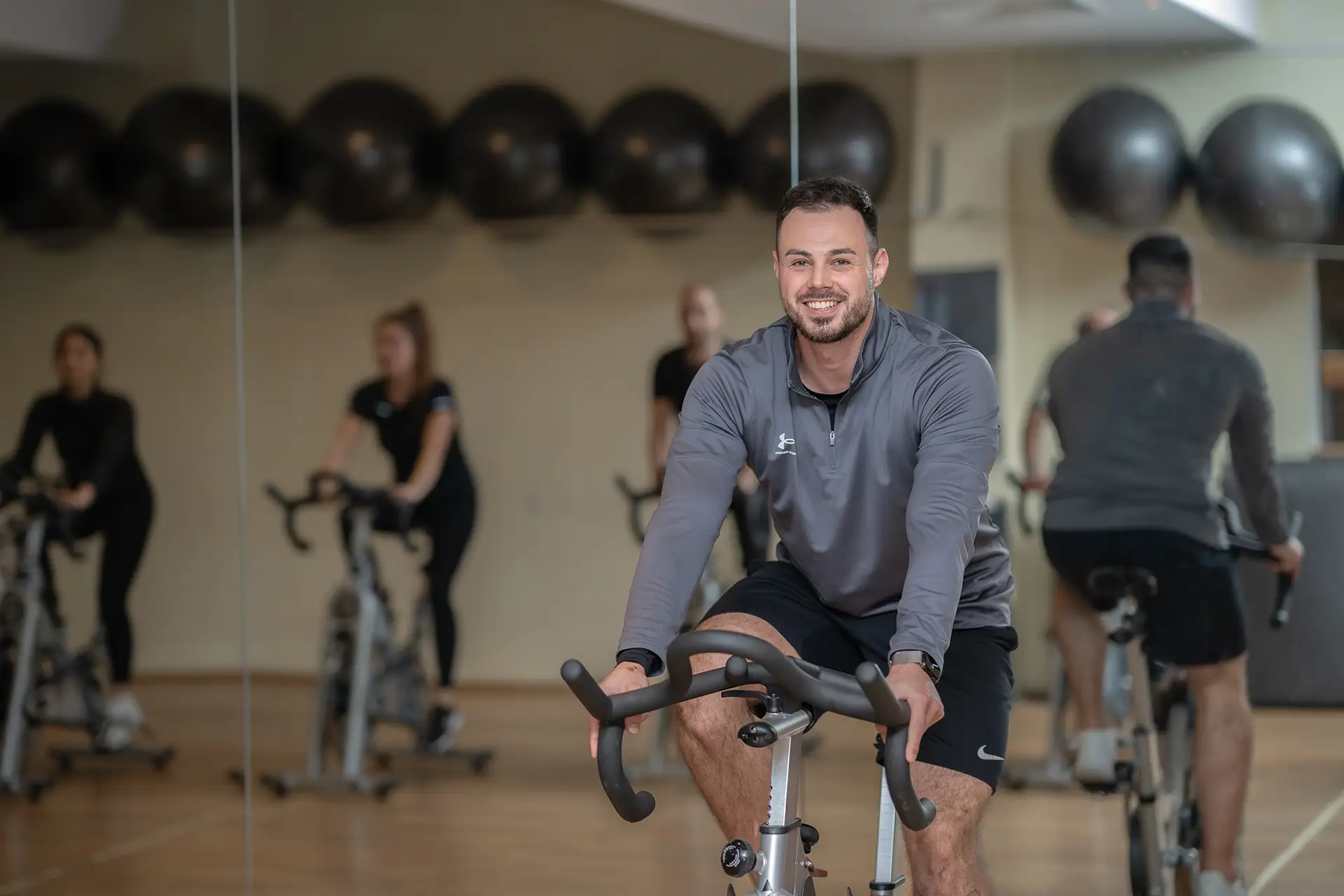 Mann trainiert lächelnd auf einem Indoor-Bike in einem Gruppenkurs im Fitnessstudio body & soul Billerbeck, weitere Teilnehmende sind im Spiegel im Hintergrund zu sehen