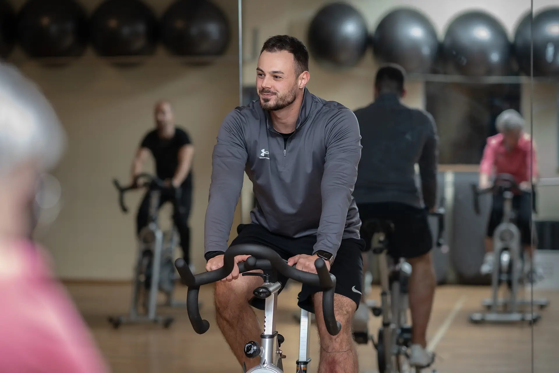 Trainer auf einem Indoor-Cycling-Bike in einem hellen Kursraum, der lächelnd nach vorne schaut. Im Hintergrund sind weitere Teilnehmende auf Fahrrädern sowie eine Spiegelwand und Gymnastikbälle zu sehen. Die Atmosphäre wirkt ruhig, motivierend und fokussiert.
