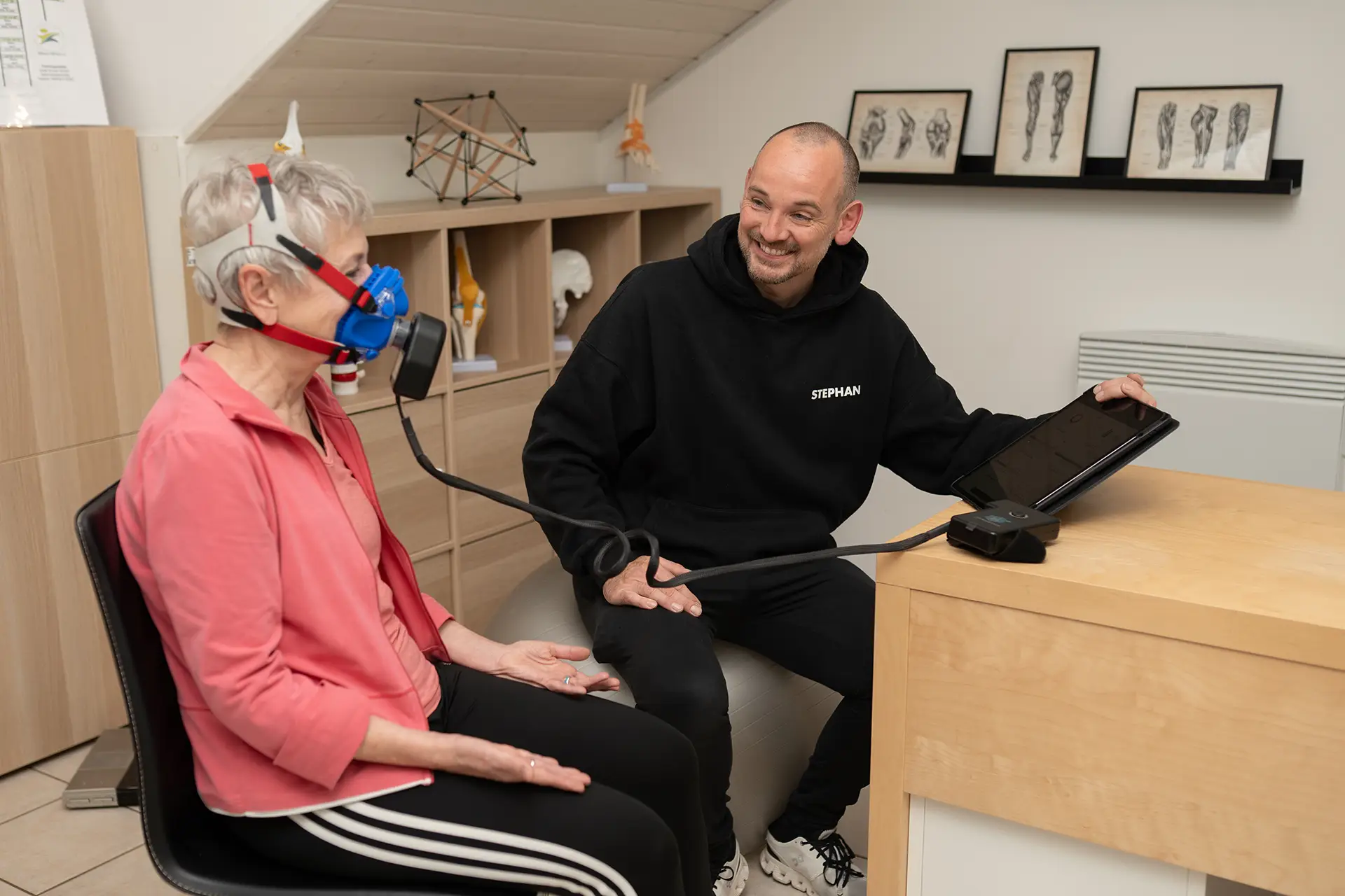 Trainer erklärt einer älteren Kundin mit Atemmaske die Ergebnisse einer Leistungs- und Stoffwechselanalyse am Tablet in einem hellen Beratungsraum im Fitnessstudio.
