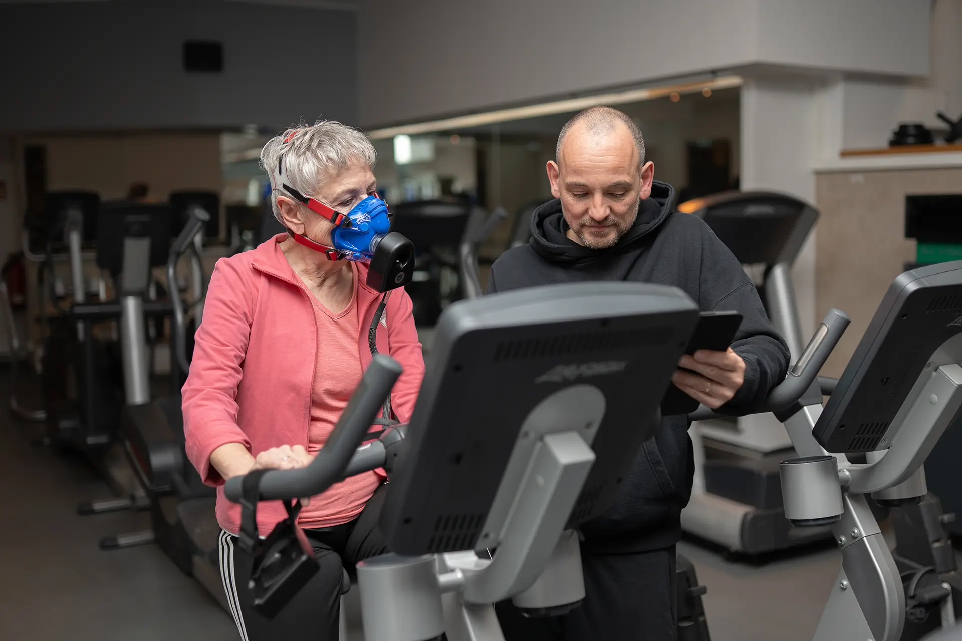 Trainer begleitet eine Kundin auf dem Crosstrainer und wertet parallel ihre Trainings- und Leistungsdaten auf einem Tablet aus, während sie mit Atemmaske trainiert.