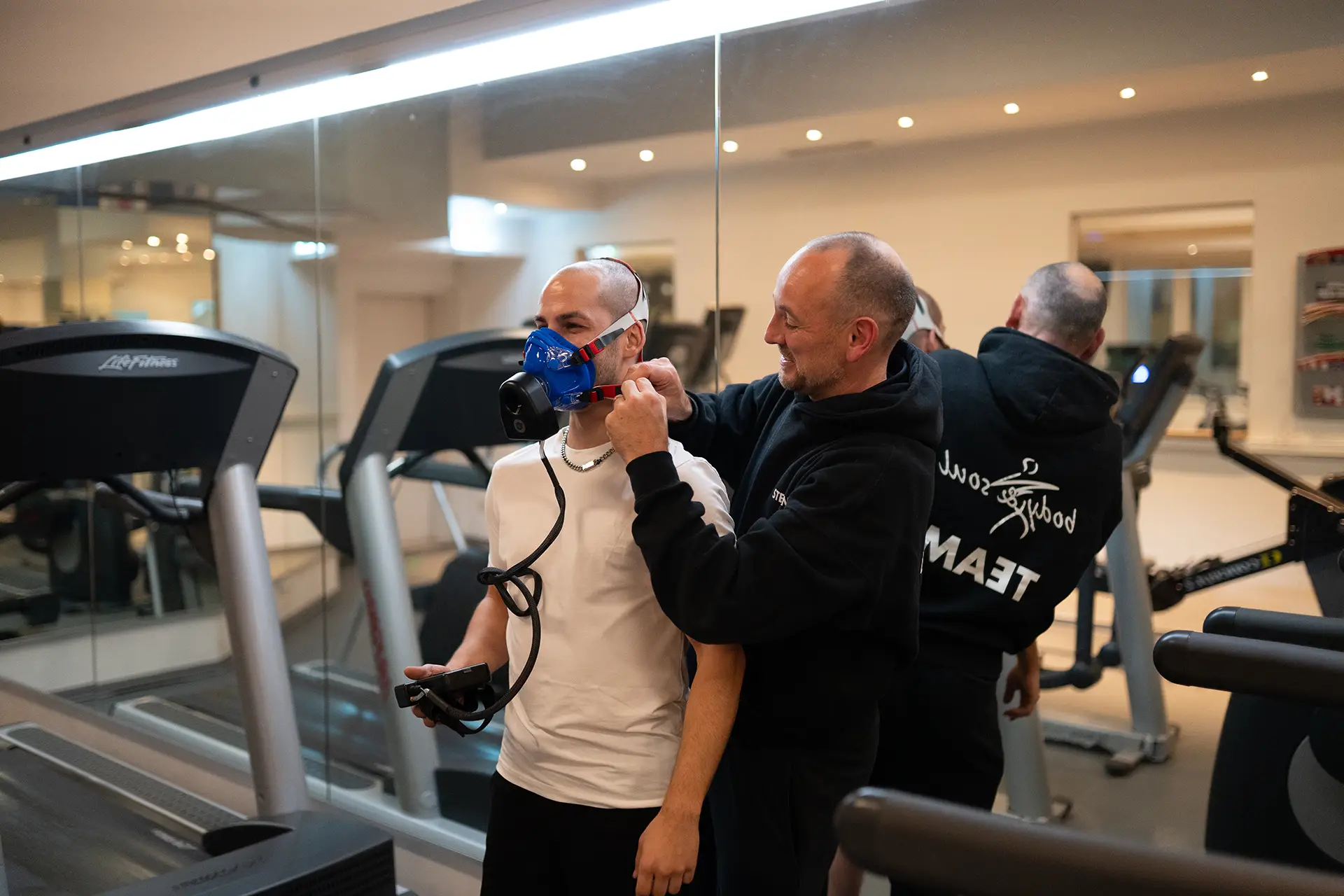 Trainer legt einem Kunden auf dem Laufband eine Atemmaske an und erklärt die Anwendung, während beide sich im Spiegel des Fitnessstudios sehen. Der Kunde hält das Messgerät in der Hand und bereitet sich auf das leistungsdiagnostische Training vor.