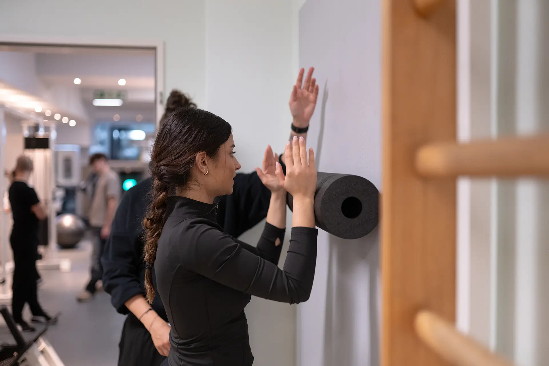 Mitglied führt eine Mobilisationsübung an der Wand im Fitnessstudio Body & Soul in Billerbeck durch, während ein Trainer im Hintergrund die Armposition und Haltung anleitet. Ein Schaumstoffroller ist an der Wand befestigt, weitere Trainierende sind im Raum zu sehen.