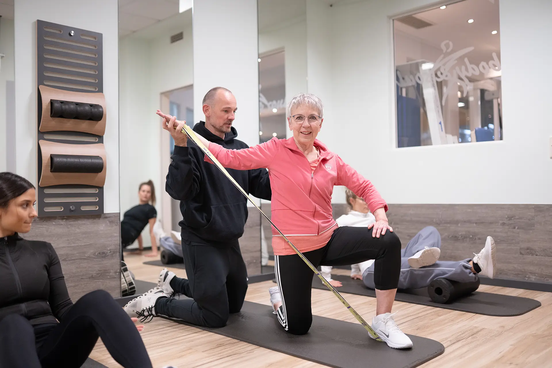 Mitglied trainiert mit einem Widerstandsband im Fitnessstudio Body & Soul in Billerbeck, während ein Trainer kniend die Übung anleitet und die Haltung kontrolliert. Im Hintergrund sitzen weitere Personen auf Trainingsmatten und führen Bodenübungen aus.