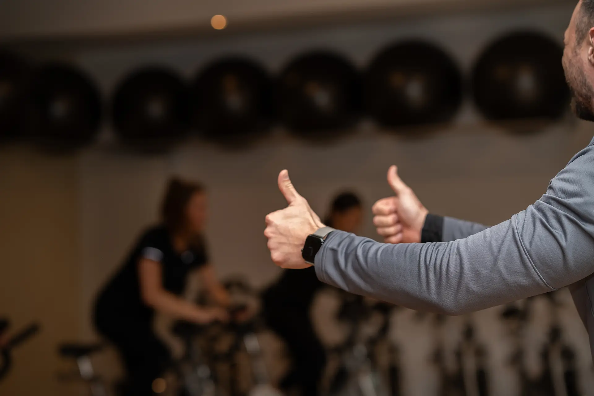 Trainer im Vordergrund zeigt mit beiden Daumen nach oben, während Teilnehmende im Hintergrund auf Indoor-Bikes trainieren und sich auf die Übung konzentrieren.
