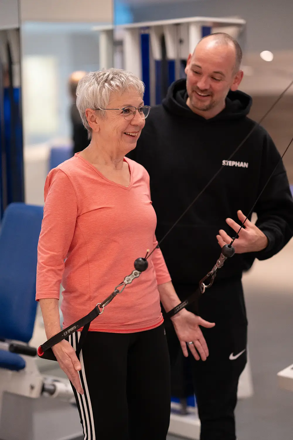 Ältere Frau trainiert an einer Seilzugmaschine im Fitnessstudio und wird von einem Trainer freundlich angeleitet und motiviert in heller, moderner Trainingsumgebung