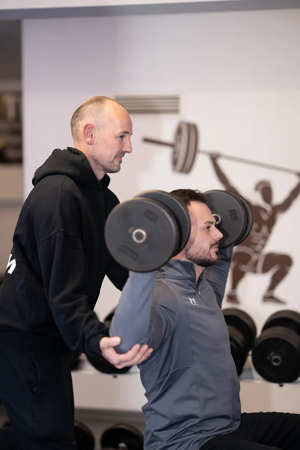 Personal Training Trainer unterstützt einen Mann beim Schulterdrücken mit Kurzhanteln im Trainingsbereich des Fitnessstudios Body & Soul in Billerbeck, im Hintergrund sind Hantelablagen und eine Wandgrafik zu sehen.