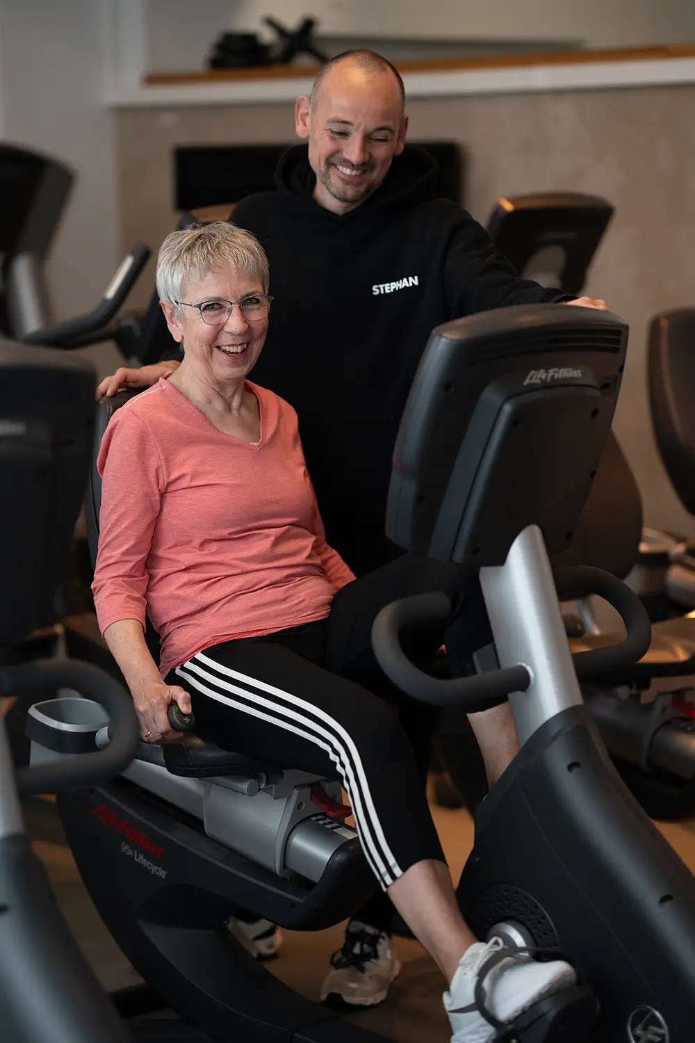 Trainer begleitet eine ältere Frau beim Training auf einem Sitz-Ergometer im Fitnessstudio body & soul Billerbeck und steht unterstützend neben dem Gerät