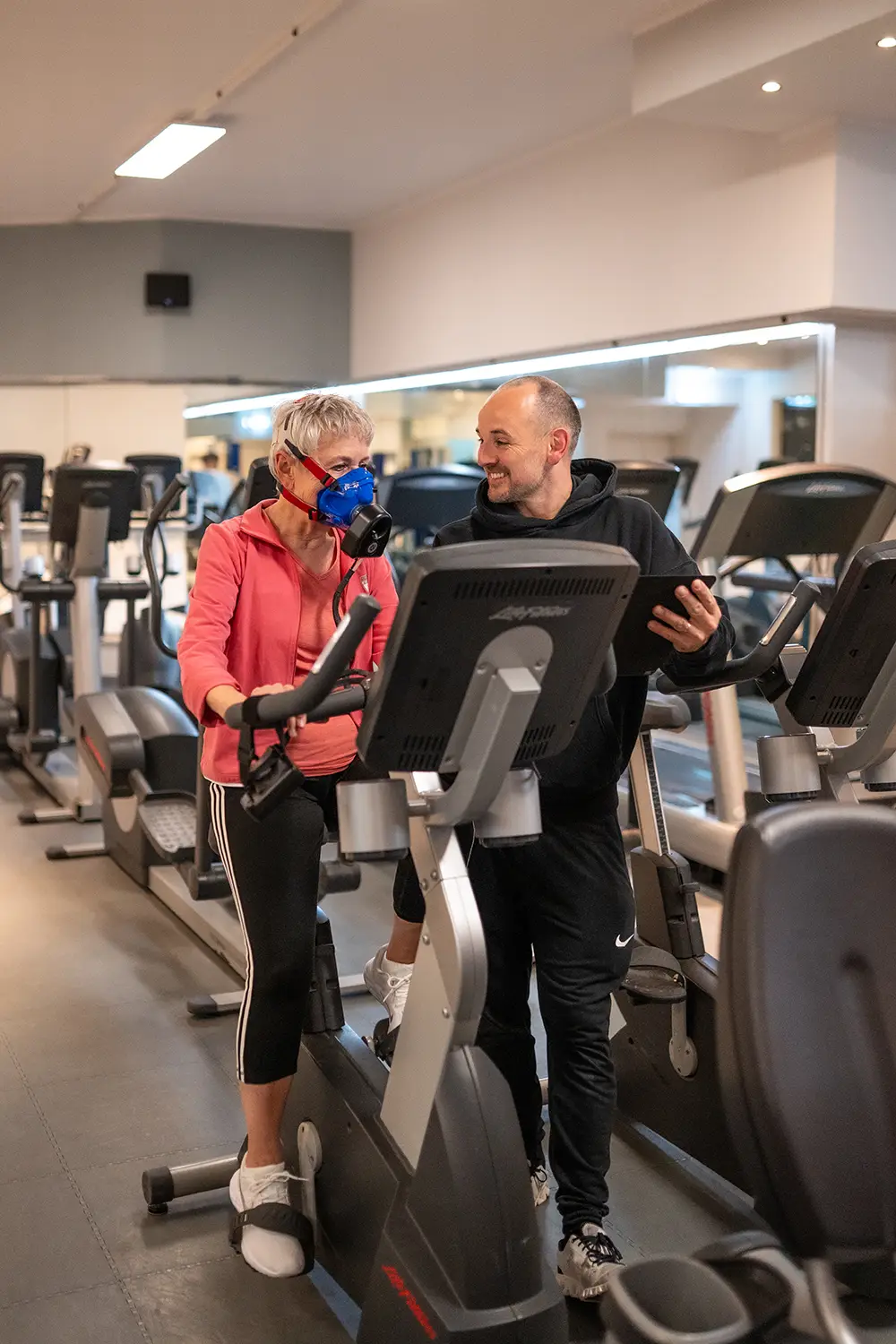Mitglied trainiert auf einem Cardiogerät im Fitnessstudio Body & Soul in Billerbeck und trägt eine Atemmaske zur Leistungsdiagnostik, während ein Trainer daneben steht und die Werte auf einem Tablet erklärt und begleitet.