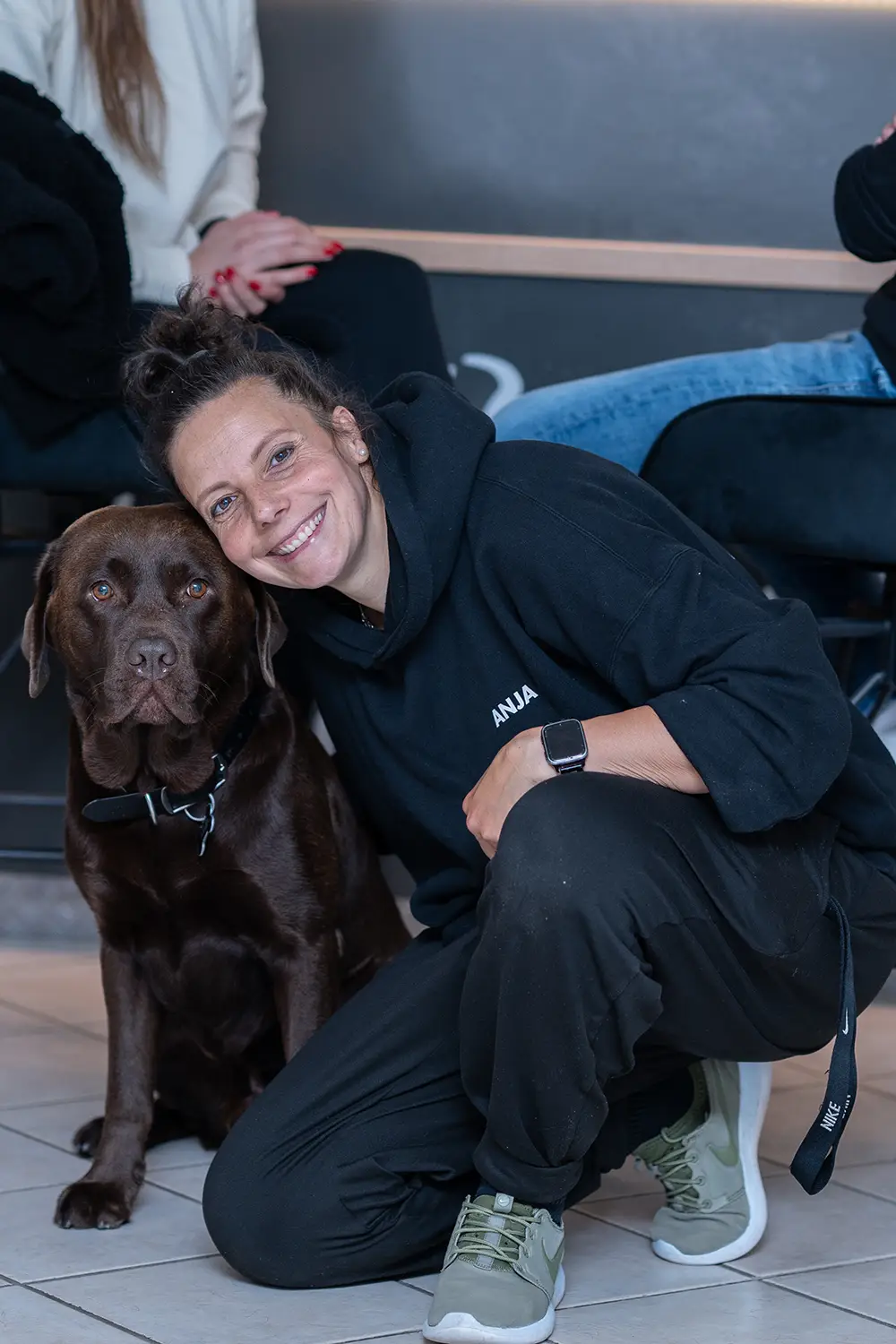 Team & Studiohund Mitarbeiterin kniet lächelnd neben einem braunen Studiohund im Empfangsbereich des Fitnessstudios Body & Soul in Billerbeck, im Hintergrund sitzen Mitglieder.